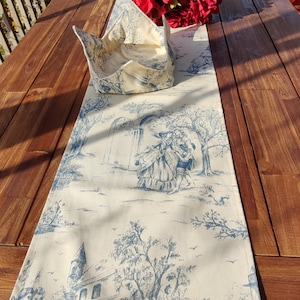 May include: A white table runner with a blue toile pattern featuring a house, trees, and people. The runner is on a wooden table with a bouquet of red flowers.