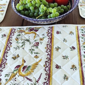 May include: A quilted placemat with a bird and floral design, featuring yellow, green, and purple accents. A bowl of tomatoes and green grapes sits on a wooden table, alongside other placemats.
