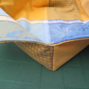 Op de afbeelding: Een gevouwen gele en blauwe stoffen broodmand met een geruit patroon.