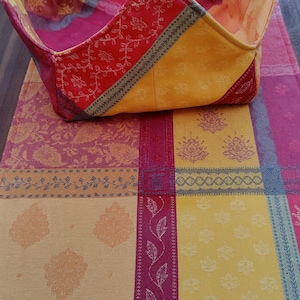 May include: A red, yellow, and orange checkered tablecloth with a floral pattern. A matching fabric basket is folded on top of the tablecloth. The basket is made of fabric and has a yellow flower sewn on the front.