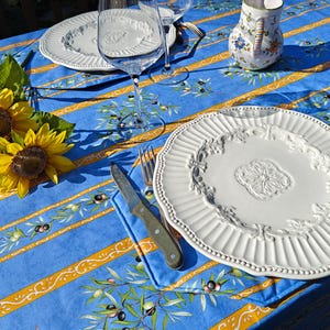 May include: A table setting with a blue tablecloth featuring olive branch designs and yellow stripes. White plates, wine glasses, a small pitcher with floral accents, and silverware are arranged on the table. Sunflowers add a touch of color.