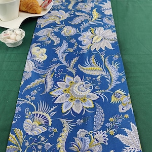 May include: A blue table runner with a floral pattern in shades of yellow, white, and grey. The runner is on a green tablecloth.