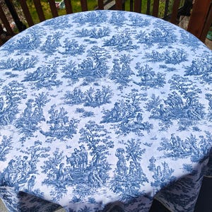 May include: A round tablecloth with a blue and white toile pattern. The design features detailed illustrations of people, trees, and buildings. The tablecloth is draped over a round table, with the edge of the table visible.