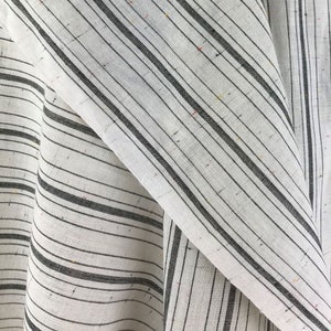 May include: Close-up of a white fabric with thin black stripes. The fabric has a subtle speckled texture.