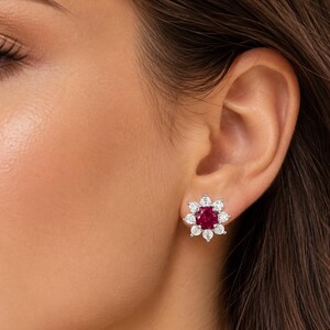 May include: A stud earring featuring a central round-cut deep red gemstone surrounded by a halo of eight small, clear, round-cut white stones arranged in a floral pattern, set in a polished silver-toned metal base.