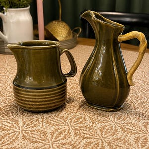 May include: Two olive green ceramic pitchers. The left pitcher has a wide, rounded base with horizontal ribbed detailing. The right pitcher features a taller, slender body with a curved, light-colored handle resembling wood grain texture.