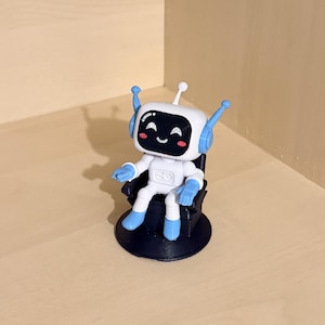May include: A small white and light blue robot figurine with a black face screen featuring a smiling expression, pink cheeks, and two antennae. The robot is seated in a black chair with a circular base, constructed with visible 3D printed layers.