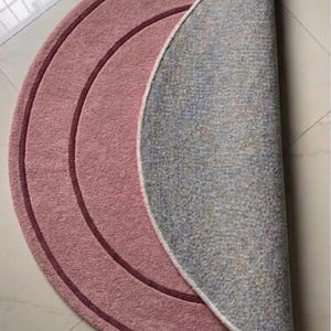 May include: A circular rug folded in half, featuring a dusty rose pink top surface with two concentric dark maroon circular lines. The underside of the rug is a light gray textured material. The text TheHandRugsCo is printed in the bottom corner.