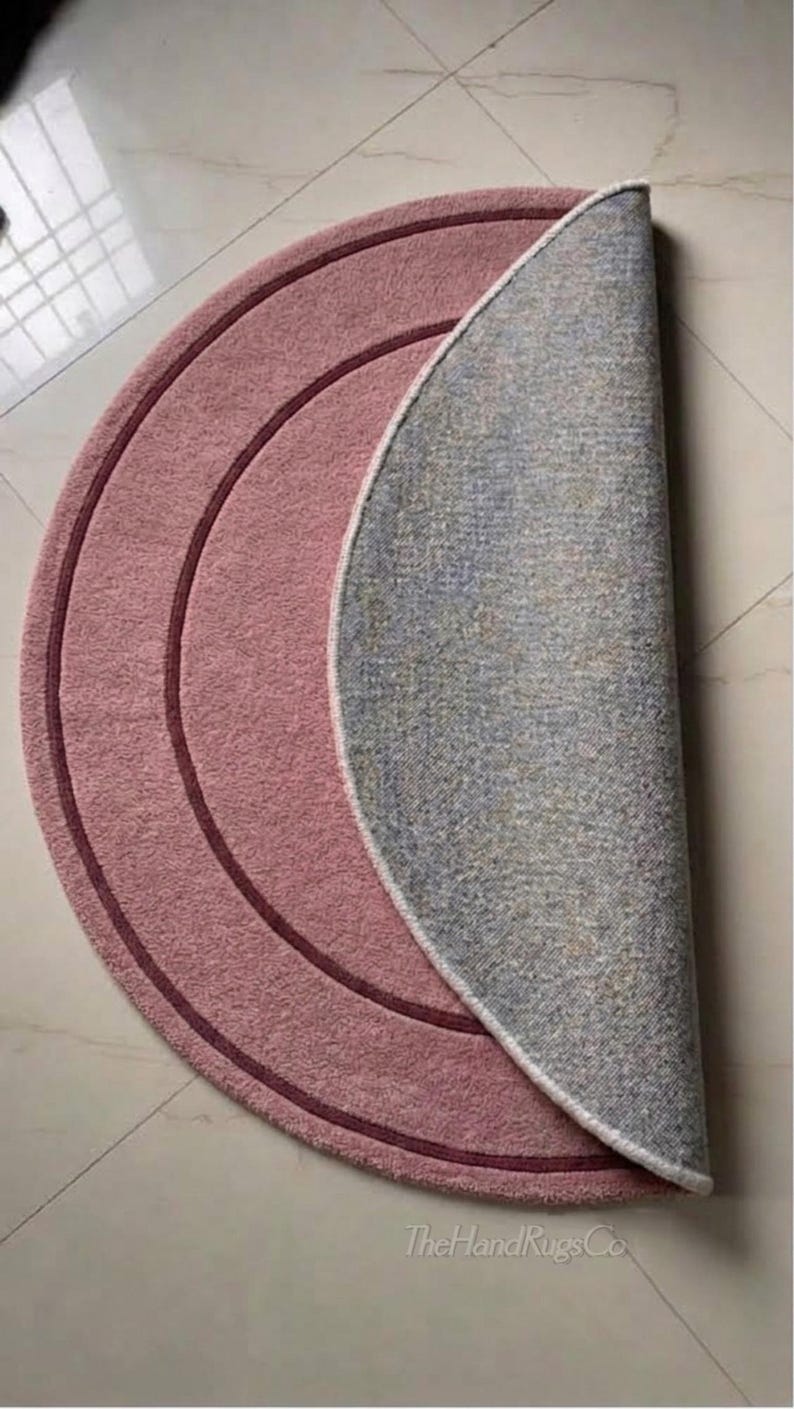 May include: A circular rug folded in half, featuring a dusty rose pink top surface with two concentric dark maroon circular lines. The underside of the rug is a light gray textured material. The text TheHandRugsCo is printed in the bottom corner.