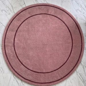 May include: A circular rug in a dusty rose color featuring a darker maroon circular border accent. The rug has a soft, textured surface and includes the text TheHandRugsCo printed in a small, light font at the bottom center of the piece.