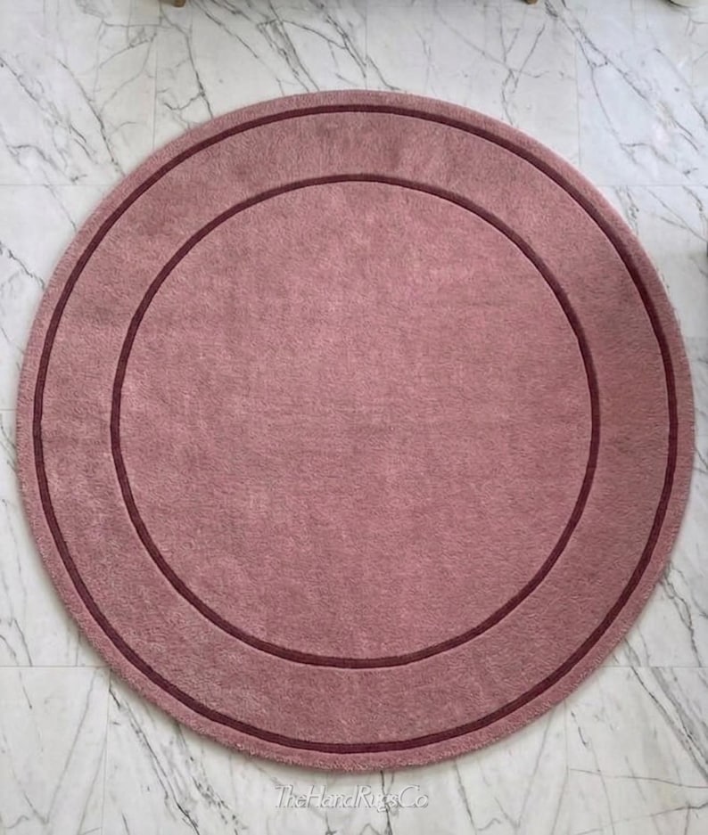 May include: A circular rug in a dusty rose color featuring a darker maroon circular border accent. The rug has a soft, textured surface and includes the text TheHandRugsCo printed in a small, light font at the bottom center of the piece.