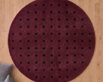 May include: A circular deep burgundy rug featuring a repeating grid pattern of small, evenly spaced black square cutouts. The rug has a plush, textured surface and is centered on a light-toned wooden floor with two gray pillows visible.