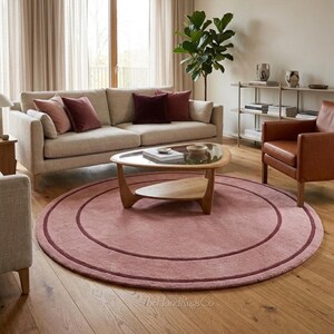 May include: A large, circular, dusty pink area rug featuring a darker maroon concentric circle design. The rug has a soft, textured pile and is positioned on a light-colored wooden floor. The text TheHandRugsCo is visible at the bottom.