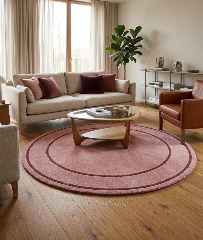 May include: A large, circular, dusty pink area rug featuring a darker maroon concentric circle design. The rug has a soft, textured pile and is positioned on a light-colored wooden floor. The text TheHandRugsCo is visible at the bottom.