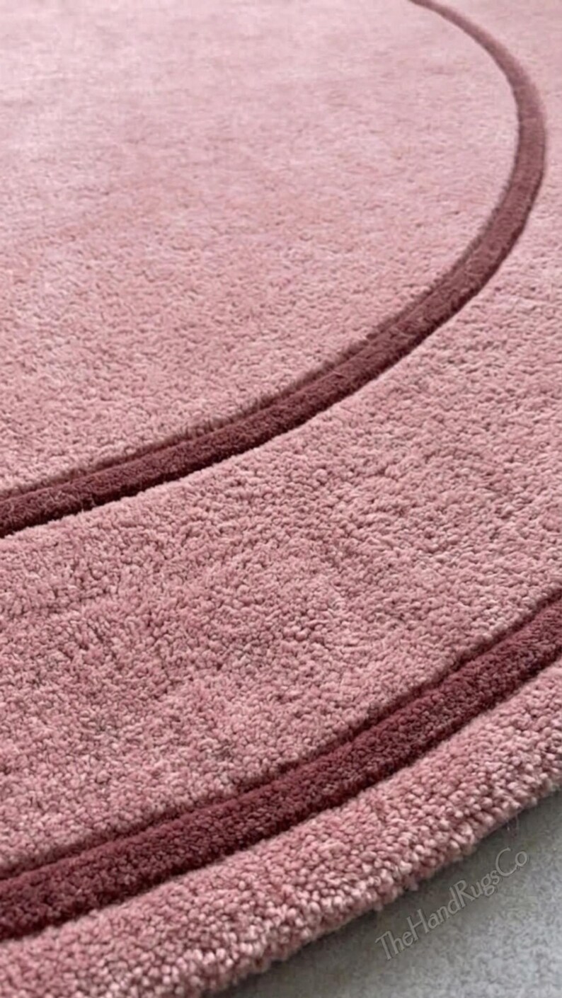 May include: A circular rug with a textured, light pink pile surface featuring a darker, deep pink circular border line. The text TheHandRugsCo is printed in the bottom right corner of the rug in a simple, light-colored font.
