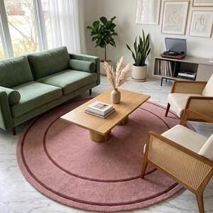 May include: A large, circular, dusty rose area rug featuring a darker pink concentric ring design. The rug is positioned on a white marble floor, serving as a central foundation for a wooden coffee table, a green sofa, and two cane chairs.