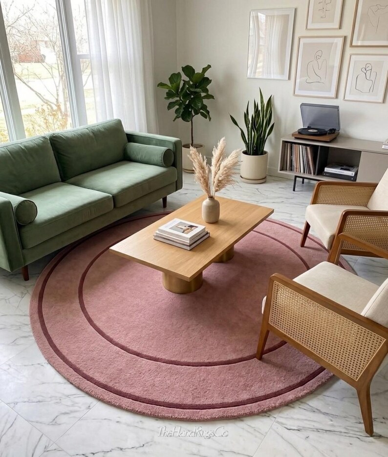 May include: A large, circular, dusty rose area rug featuring a darker pink concentric ring design. The rug is positioned on a white marble floor, serving as a central foundation for a wooden coffee table, a green sofa, and two cane chairs.