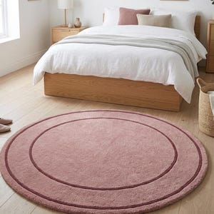 May include: A circular, dusty rose-colored area rug featuring a darker, thin circular border accent. The rug has a soft, textured pile surface and is positioned on a light-colored wooden floor. The text TheHandRugsCo is visible.