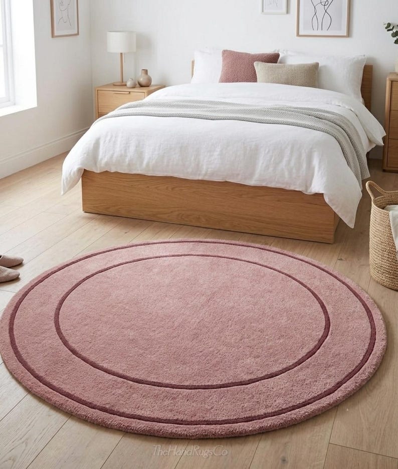 May include: A circular, dusty rose-colored area rug featuring a darker, thin circular border accent. The rug has a soft, textured pile surface and is positioned on a light-colored wooden floor. The text TheHandRugsCo is visible.