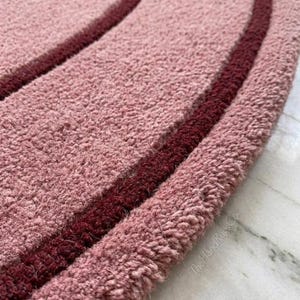 May include: A circular rug with a thick, tufted texture in a dusty rose color, featuring two concentric rings in a deep burgundy shade. The text TheHandRugsCo is visible in small, light-colored lettering near the edge of the rug's surface.