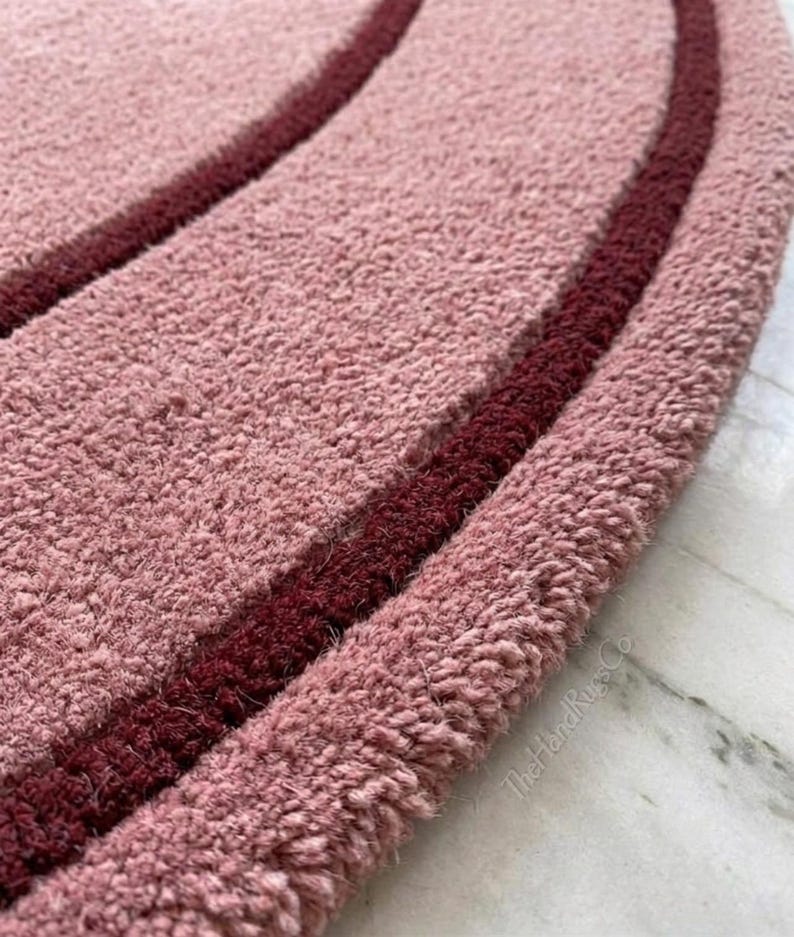 May include: A circular rug with a thick, tufted texture in a dusty rose color, featuring two concentric rings in a deep burgundy shade. The text TheHandRugsCo is visible in small, light-colored lettering near the edge of the rug's surface.