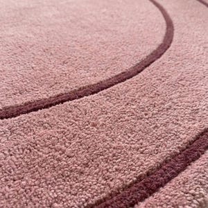 May include: A circular rug with a textured, light pink pile surface featuring two concentric rings in a darker, deep burgundy shade. The rug has a dense, tufted appearance and includes the text TheHandRugsCo in the bottom corner.