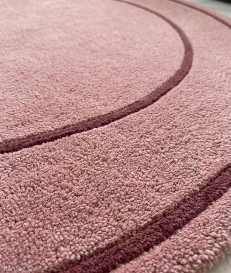 May include: A circular rug with a textured, light pink pile surface featuring two concentric rings in a darker, deep burgundy shade. The rug has a dense, tufted appearance and includes the text TheHandRugsCo in the bottom corner.