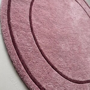 May include: A circular rug in a dusty rose color featuring a darker maroon circular border accent. The rug has a textured, plush pile surface and includes the text TheHandRugsCo printed in a light color near the bottom edge.