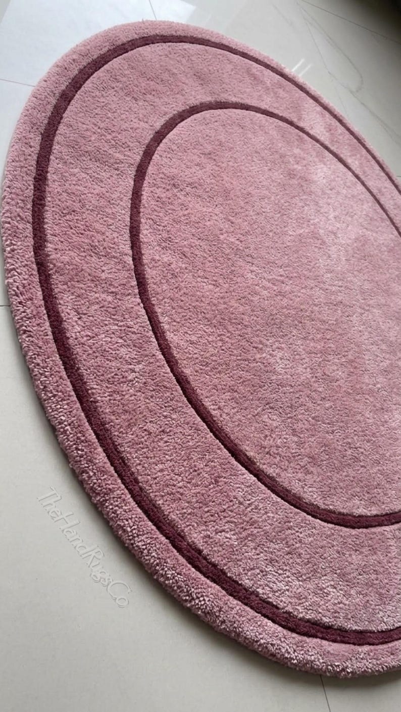 May include: A circular rug in a dusty rose color featuring a darker maroon circular border accent. The rug has a textured, plush pile surface and includes the text TheHandRugsCo printed in a light color near the bottom edge.