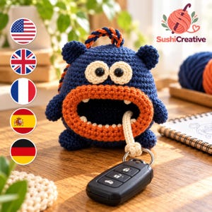 May include: A dark blue crocheted monster keychain pouch with a large orange mouth, two cream-colored eyes with black pupils, and a braided orange and blue handle. A black car key fob is attached to the pouch by a cream-colored cord.