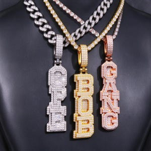May include: Three vertical pendant necklaces featuring iced-out block letters. The left pendant spells OPE, the center spells BOB, and the right spells GANG. Each piece is encrusted with small clear stones and hangs from a thick metal chain.