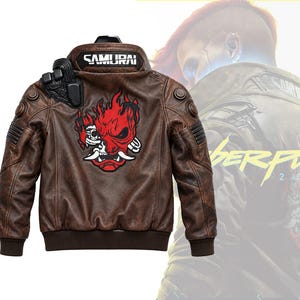 Könnte beinhalten: Eine braune Lederjacke mit einer großen rot-schwarzen Grafik einer stilisierten Dämonenmaske auf dem Rücken. Am Kragen steht in weißen Buchstaben SAMURAI. Die Jacke hat schwarze Schulterakzente sowie gerippte Ärmelbündchen und einen gerippten Saum.