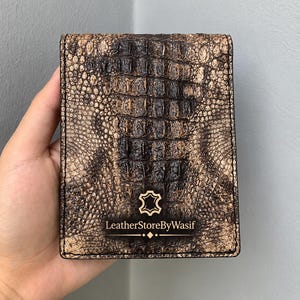 Könnte beinhalten: Ein rechteckiges Klappportemonnaie mit einer strukturierten Krokodilleder-Optik in Braun und Schwarz. Auf der Vorderseite befindet sich ein Logo mit einem Lederhaut-Symbol und dem Text LeatherStoreByWasif in serifenloser Schrift.