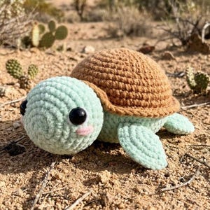 May include: A crocheted turtle toy featuring a light mint green head and flippers, a textured brown shell, large black plastic safety eyes, and small pink stitched cheeks. The stuffed animal has a rounded, soft yarn construction.