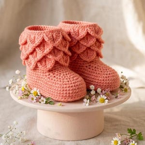 May include: A pair of peach-colored crochet booties featuring a textured, layered leaf-like pattern around the ankle cuffs. The booties are crafted with a dense stitch and have a rounded toe design, showcasing a soft, uniform yarn texture throughout.