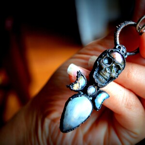 May include: A silver pendant featuring a black skull with iridescent accents, surrounded by a cluster of iridescent stones and a teardrop-shaped blue stone.