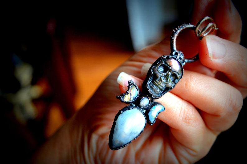 May include: A silver pendant featuring a black skull with iridescent accents, surrounded by a cluster of iridescent stones and a teardrop-shaped blue stone.