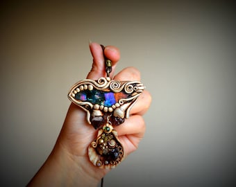 May include: A handmade pendant featuring a butterfly-shaped design with iridescent blue and purple accents. The pendant is made of polymer clay and includes various beads and stones, including a brown stone, a green stone, and a shell.