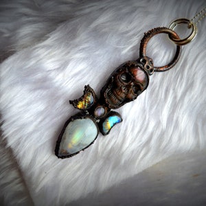 May include: A silver chain necklace with a pendant featuring a skull with a rainbow moonstone and a copper wire design.