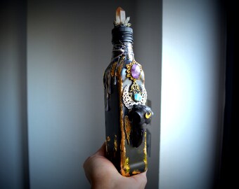 May include: A black decorative bottle with gold accents, adorned with a crystal, amethyst, and turquoise embellishments. The bottle features a sculpted owl and ornate metalwork, creating a unique, artistic design.