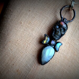 May include: A silver chain necklace with a black skull pendant. The skull is adorned with iridescent moonstone and abalone shell accents.