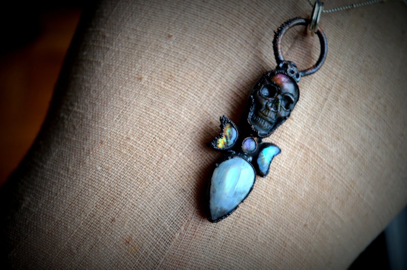 May include: A silver chain necklace with a black skull pendant. The skull is adorned with iridescent moonstone and abalone shell accents.