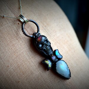 May include: A black skull pendant with iridescent gemstone accents hangs from a silver chain. The skull is detailed with a white face and black eyes. The pendant is attached to a silver ring.