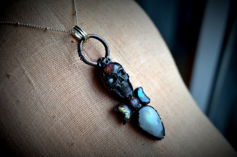 May include: A black skull pendant with iridescent gemstone accents hangs from a silver chain. The skull is detailed with a white face and black eyes. The pendant is attached to a silver ring.