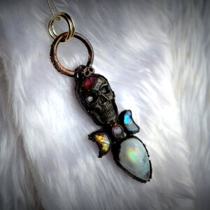 May include: A silver skull pendant necklace with a rainbow moonstone and iridescent glass accents. The pendant is on a white faux fur background.