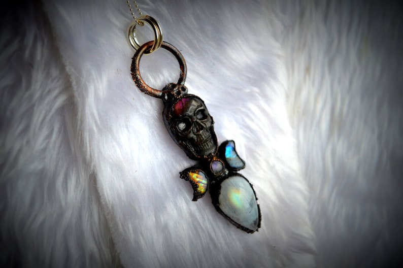 May include: A silver skull pendant necklace with a rainbow moonstone and iridescent glass accents. The pendant is on a white faux fur background.