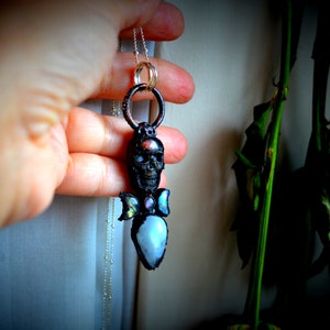 May include: A silver chain necklace with a black skull pendant. The skull has a gemstone in its forehead and two iridescent wings with gemstones on each side. The pendant is hanging from a silver ring.