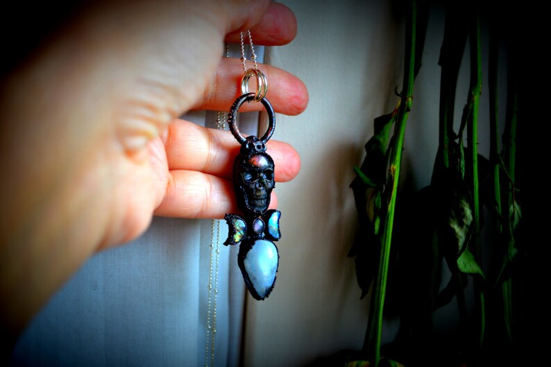 May include: A silver chain necklace with a black skull pendant. The skull has a gemstone in its forehead and two iridescent wings with gemstones on each side. The pendant is hanging from a silver ring.