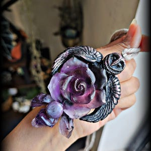 May include: A pendant featuring a central purple rose with glitter accents, flanked by dark metallic wings and a small owl head at the top. The piece includes purple crystal-like clusters at the base and hangs from a light-colored cord.
