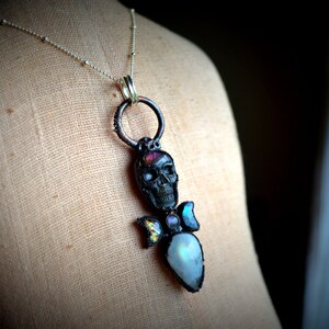 May include: A silver chain necklace with a black skull pendant accented with iridescent moonstone and a pink gemstone. The pendant is attached to a silver ring.